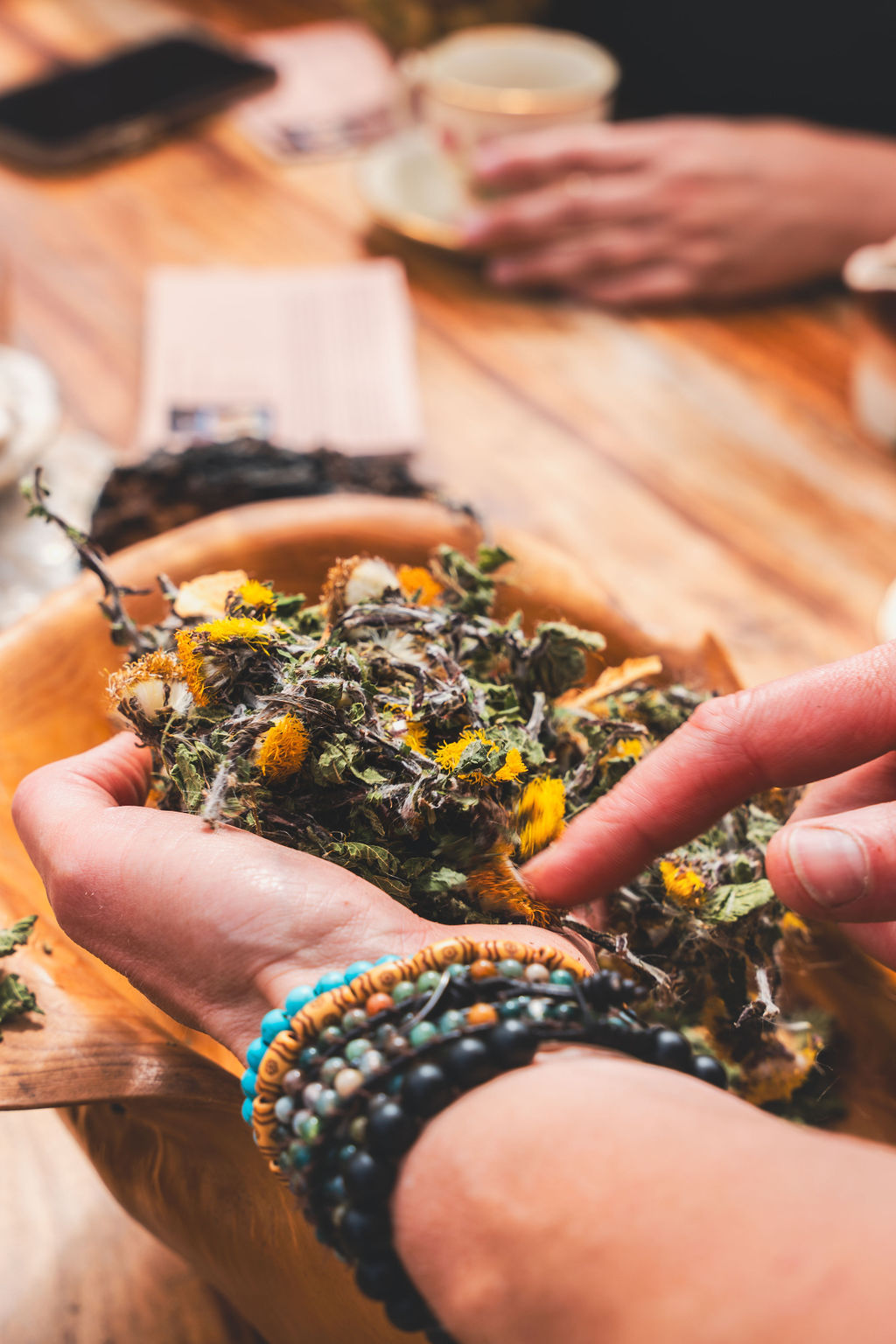 Two hands holding dried dandelion heads and leaves above a wooden bowl of foraged tea blend