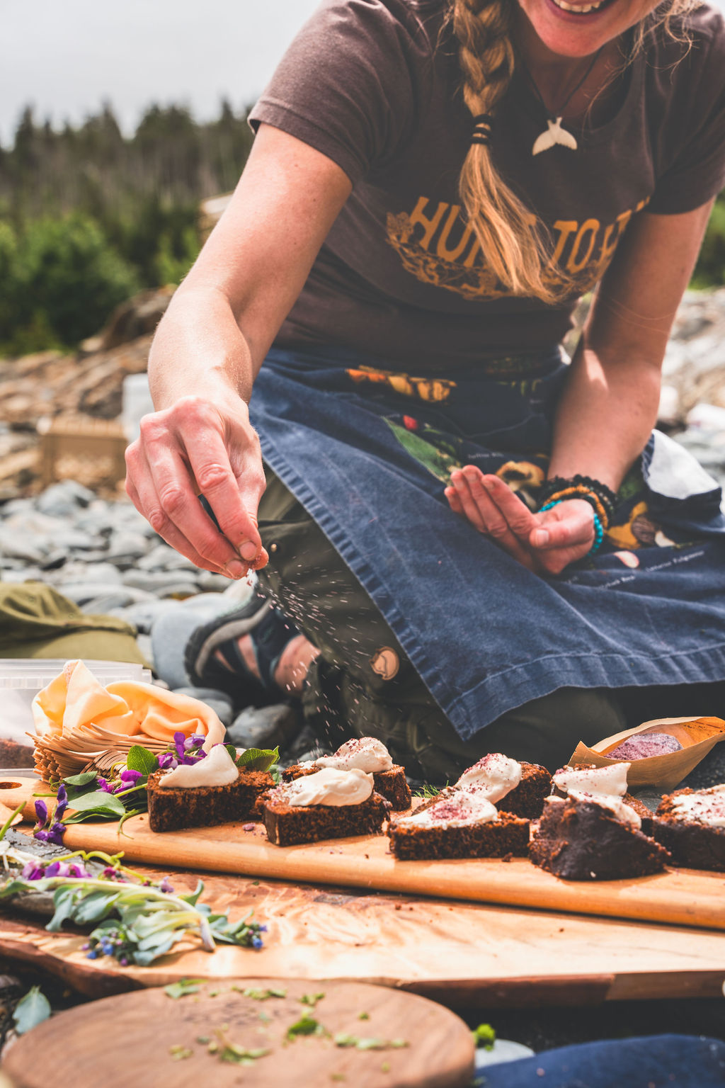 Lori McCarthy finishing dark cakes topped with cream and wild flowers on a wooden board at a shoreline cook-up